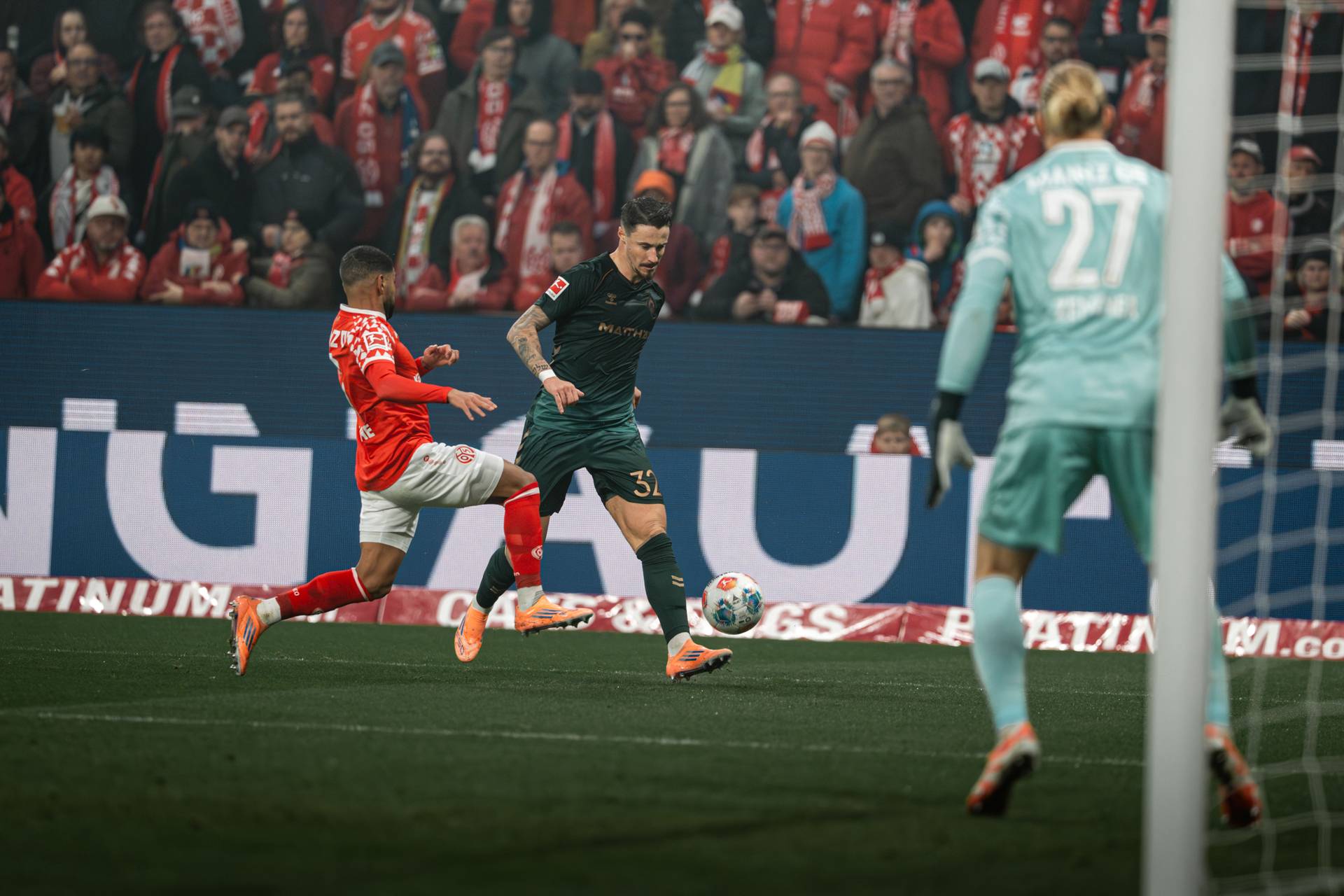 Fußballspieler in Aktion während eines Spiels, wobei ein Verteidiger in Rot versucht, einen Spieler in Grün in der Nähe des Tores zu blockieren. Ein Torwart beobachtet das Geschehen.