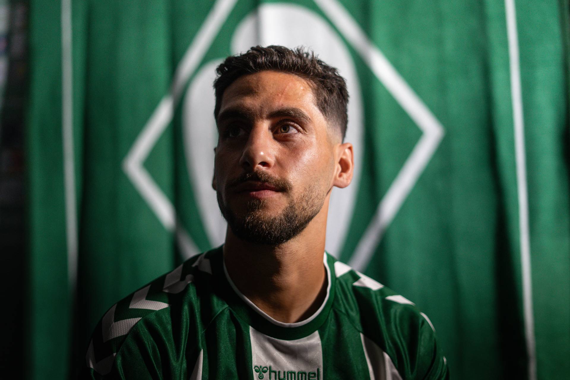 Ein Mann mit kurzen dunklen Haaren und Bart trägt ein grün-weißes Sporttrikot, blickt nach oben und steht vor einem grün-weißen Hintergrund.