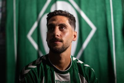 Ein Mann mit kurzen dunklen Haaren und Bart trägt ein grün-weißes Sporttrikot, blickt nach oben und steht vor einem grün-weißen Hintergrund.