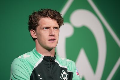 Jens Stage looks focused as he speaks, with a wall featuring the Werder badge visible in the background.