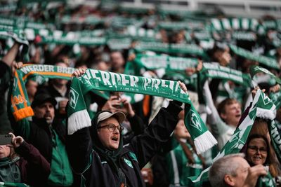 Fans, die bei einer gut besuchten Stadionveranstaltung begeistert grün-weiße Schals mit der Aufschrift „Werder Bremen“ hochhalten.
