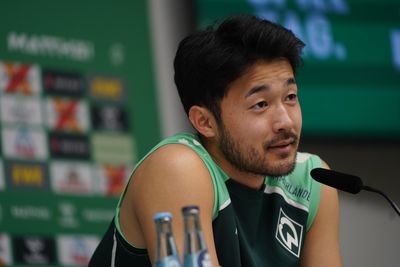 Yukinari Sugawara sitzt vor einer Werbewand am Mikrofon und lächelt.