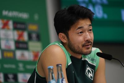 Yukinari Sugawara sitzt vor einer Werbewand am Mikrofon und lächelt.