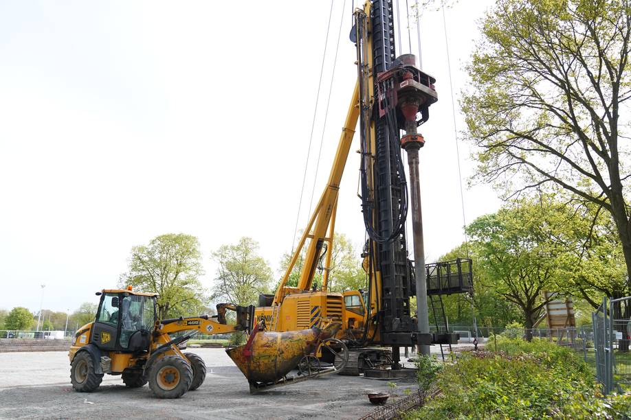Eine Baustelle mit schwerem Baumaschinenpark, darunter eine Bohranlage und ein Frontlader, vor dem Hintergrund von Bäumen und einem klaren Himmel.