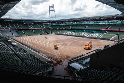 Ein Stadion, das gerade renoviert wird, mit Baufahrzeugen auf einem mit Erde bedeckten Spielfeld, umgeben von leeren grünen und weißen Sitzplätzen.