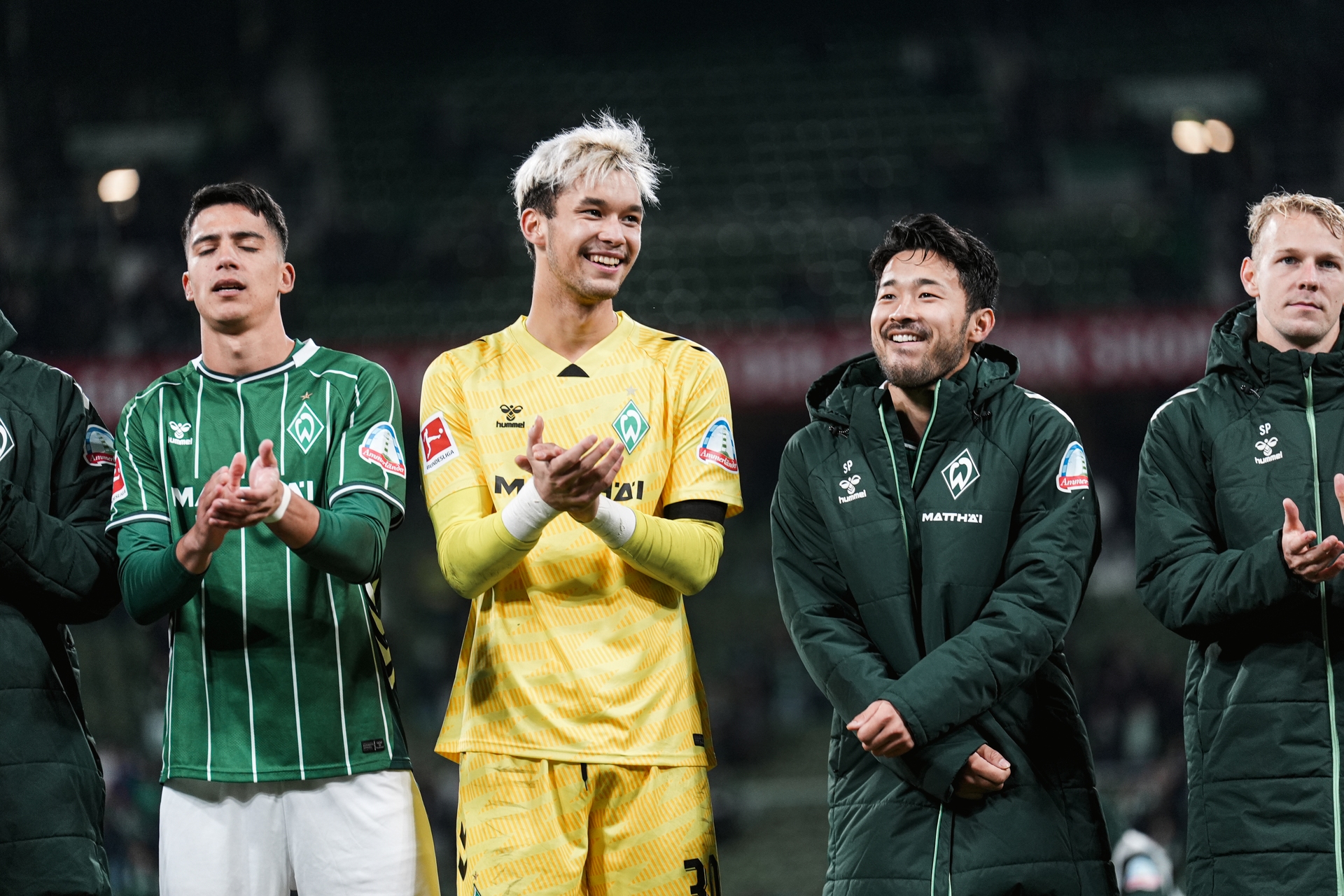 Fußballspieler stehen in einer Reihe, tragen ihre Mannschaftstrikots, klatschen und lächeln auf einem schwach beleuchteten Spielfeld.