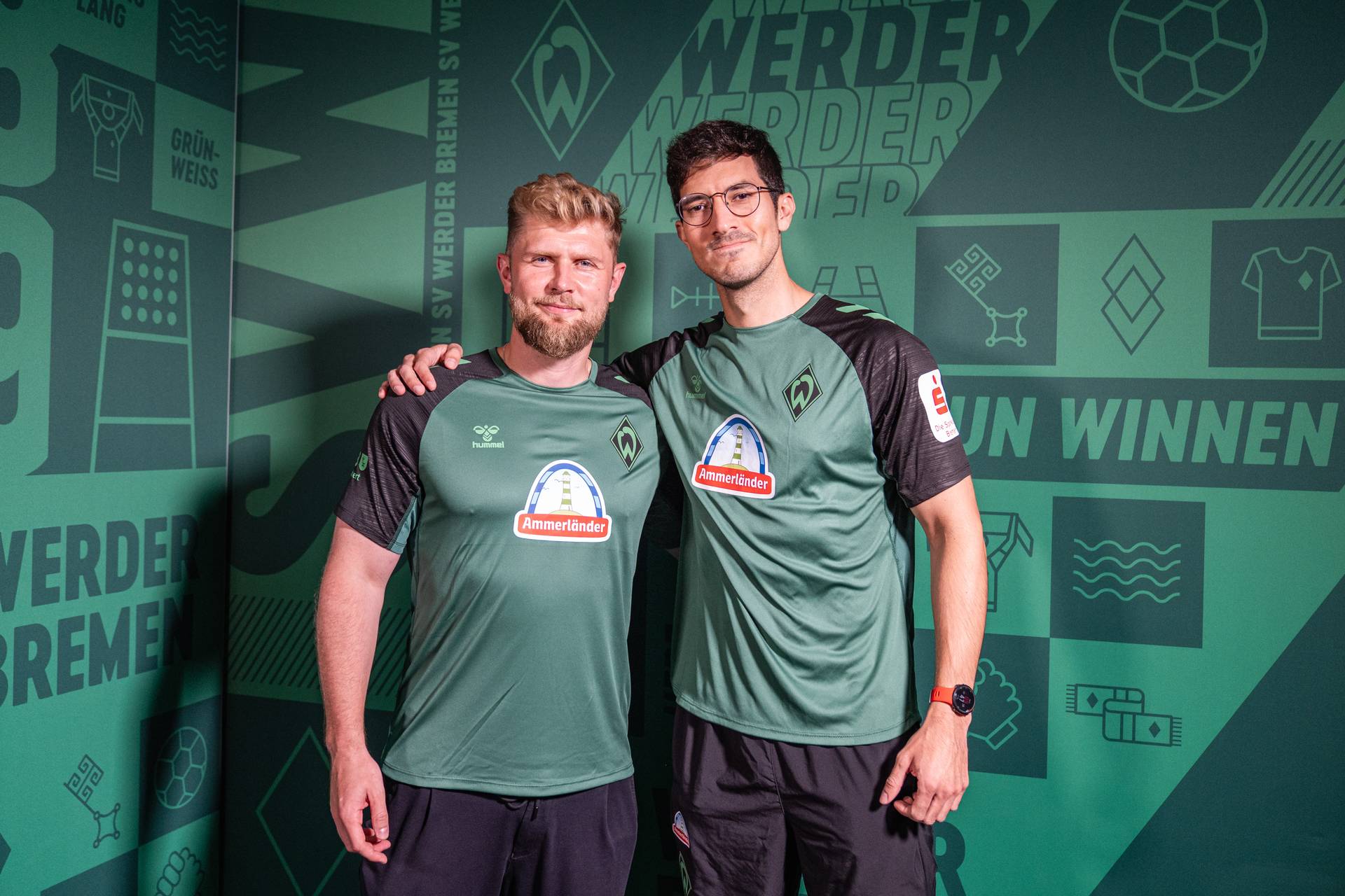 Sebastian Kmiec und Alvaro Chaparro, die Trikots von Werder Bremen tragen, stehen mit umeinander gelegten Armen vor einem grünen Hintergrund mit Vereinslogos und Schriftzügen.