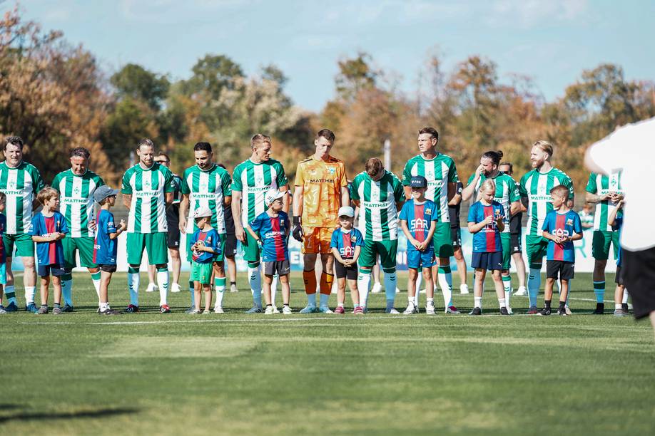 Eine Fußballmannschaft in grün gestreiften Trikots steht mit kleinen Kindern in blauen und roten Trikots auf einer Rasenfläche in einer Reihe. Im Hintergrund sind Bäume zu sehen.
