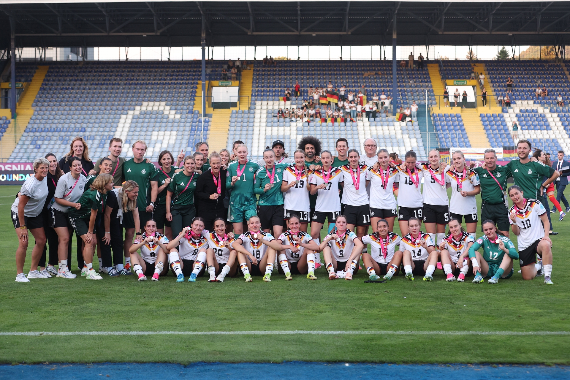 Eine Frauenfußballmannschaft posiert mit Medaillen auf dem Spielfeld vor leeren Stadionrängen und feiert ihren Erfolg.