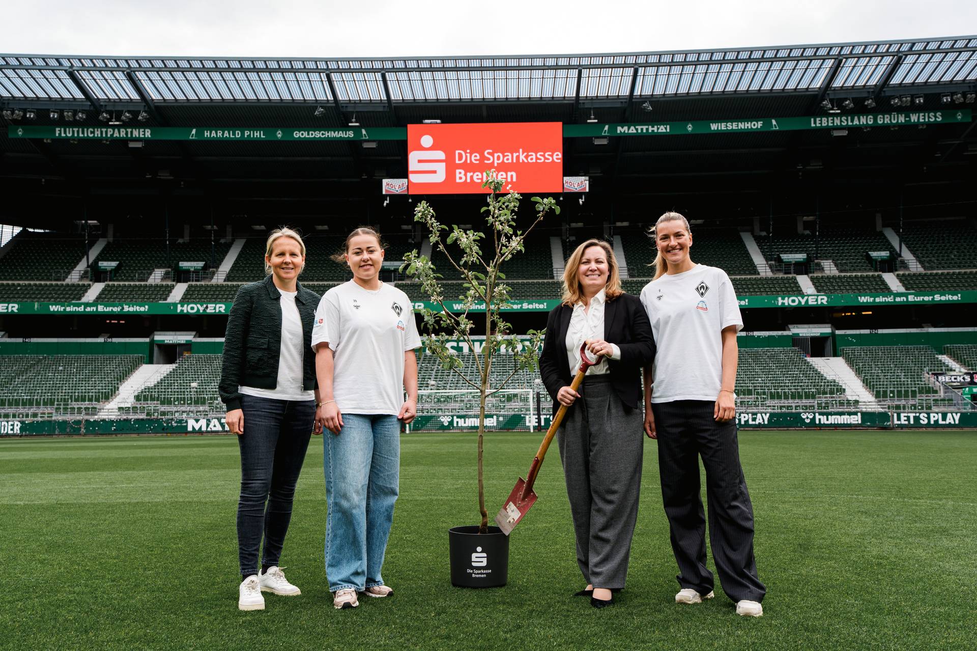 Vier Personen stehen auf einem Fußballplatz neben einem Baum. Zwei von ihnen tragen legere Sportkleidung, und im Hintergrund ist ein Schild mit der Aufschrift „Die Sparkasse Bremen“ zu sehen.