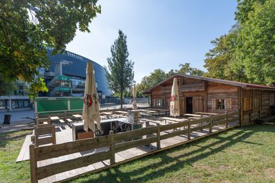 Eine hölzerne Hütte mit Sonnenschirmen und Sitzgelegenheiten, umgeben von Bäumen, in der Nähe eines modernen Glasgebäudes an einem sonnigen Tag.