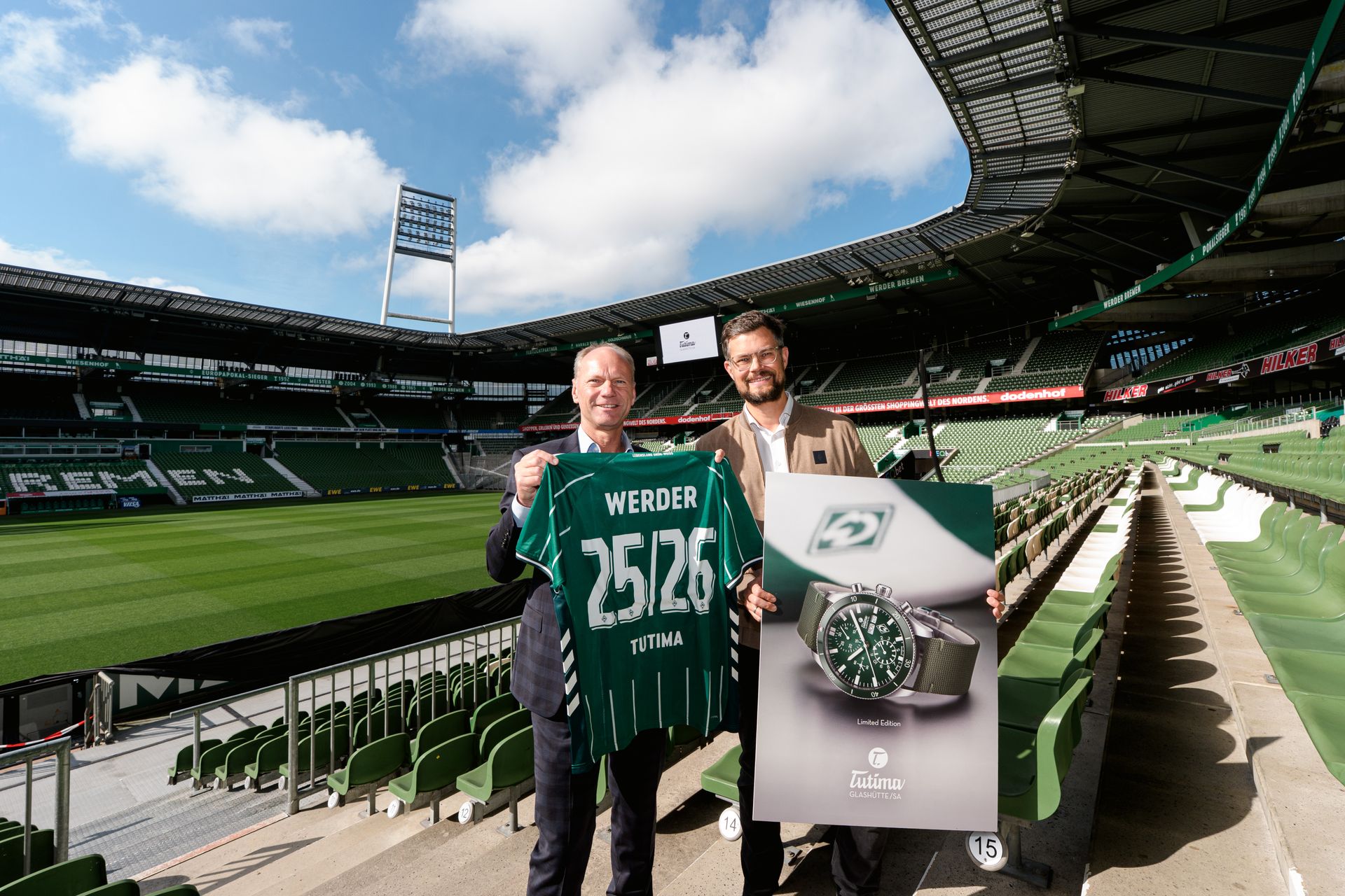 Zwei Personen stehen im Weserstadion. Sie halten ein Schild und ein Trikot. Auf dem Schild ist eine Uhr abgebildet.