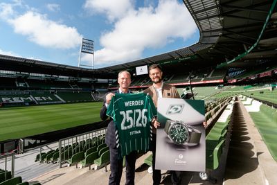 Zwei Personen stehen im Weserstadion. Sie halten ein Schild und ein Trikot. Auf dem Schild ist eine Uhr abgebildet.