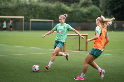Michelle Ulbrich und Isabella Jaron, die grüne Trikots tragen, trainieren auf einem Spielfeld; die eine dribbelt den Ball, während die andere zusieht. Im Hintergrund sind Tore und Grünflächen zu sehen.