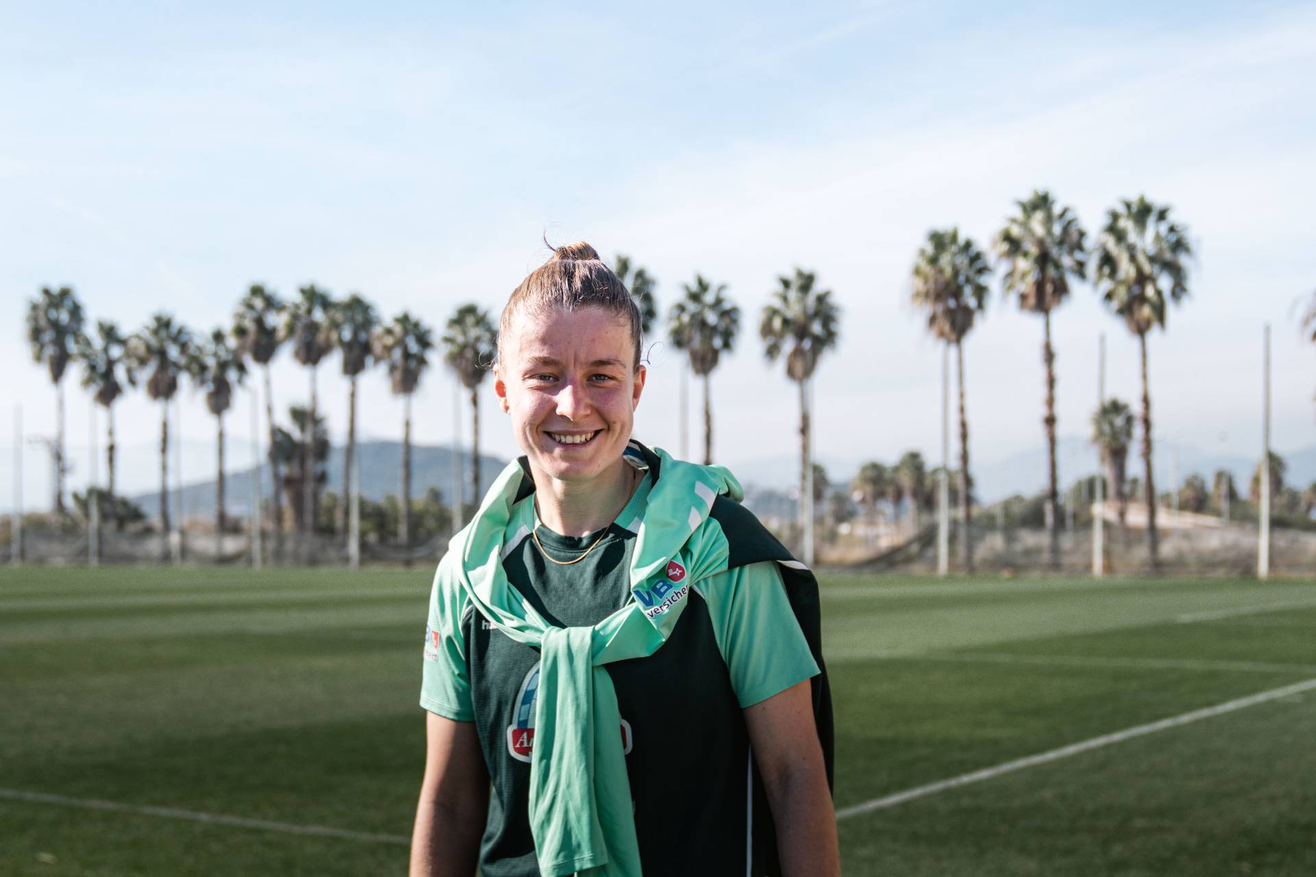 Michelle Ulbrich lächelt in die Kamera, im Hintergrund sind Palmen, ein Fußballplatz und blauer Himmel zu sehen.