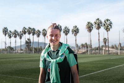 Michelle Ulbrich lächelt in die Kamera, im Hintergrund sind Palmen, ein Fußballplatz und blauer Himmel zu sehen.