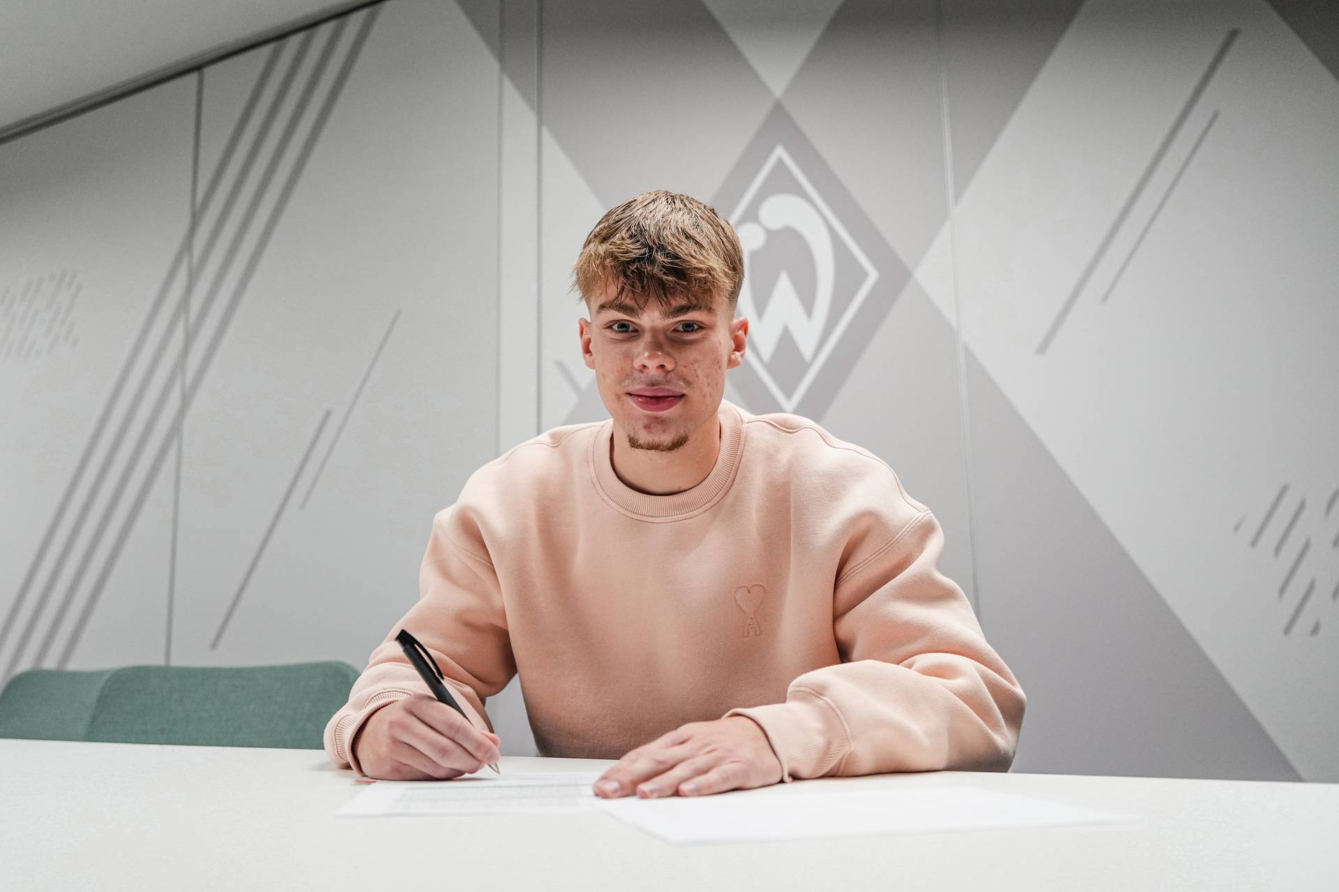 Mick Schmetgens bei der Vertragsunterschrift vor einer Wand mit der Werder-Raute.