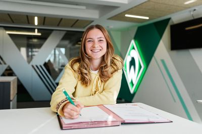 Michelle Weiß trägt ein gelbes Oberteil, lächelt in die Kamera und hält einen Stift über ihrem Vertrag.