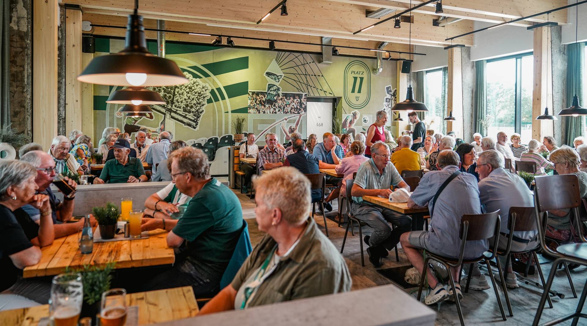 60plus Mitglieder sitzen an Holztischen im neuen Vereinsheim mit Fensterfront rechts und grüner Wand im Hintergrund