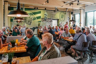 60plus Mitglieder sitzen an Holztischen im neuen Vereinsheim mit Fensterfront rechts und grüner Wand im Hintergrund