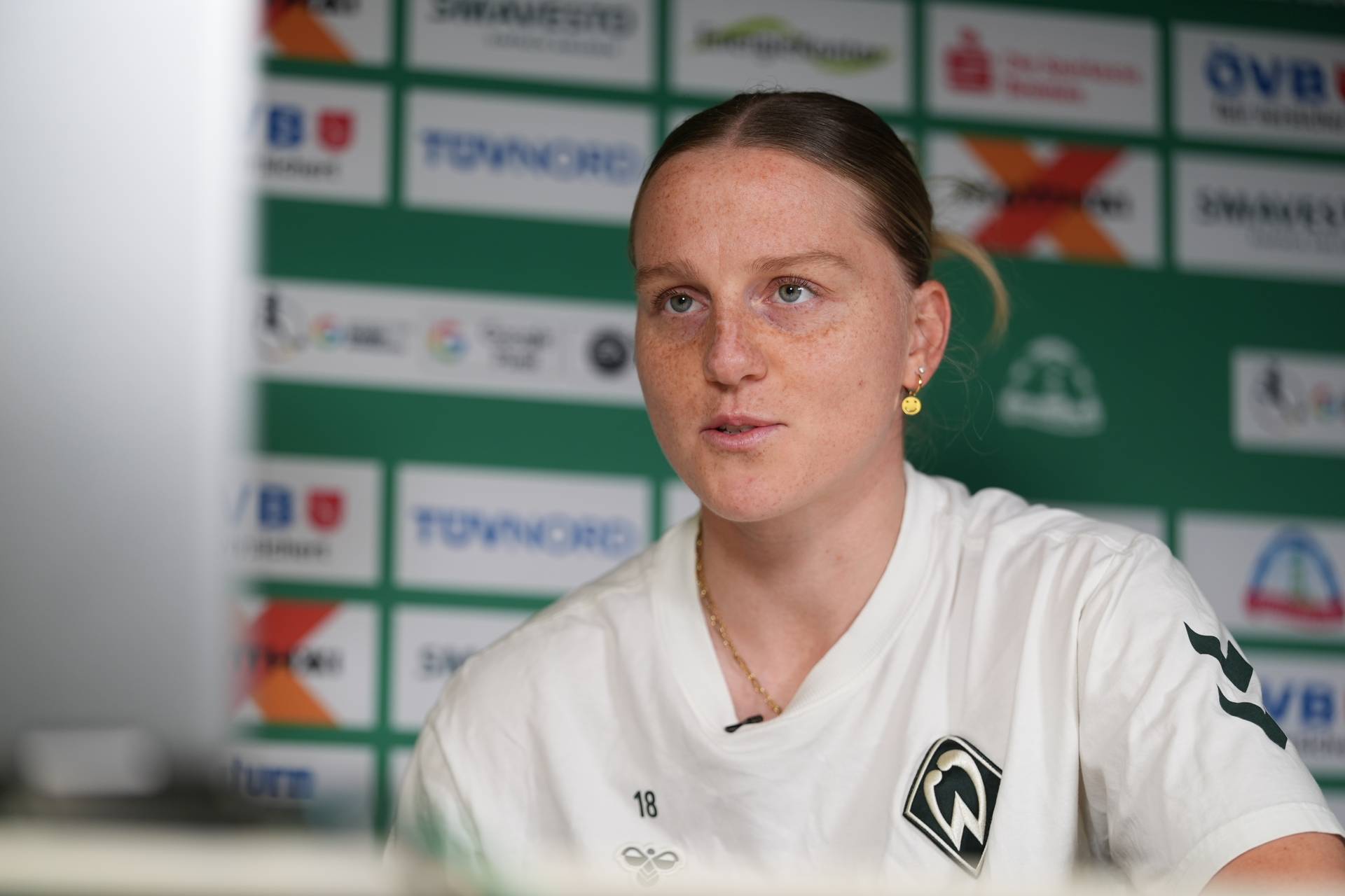Lina Hausicke sitzt im weißen Werder-Shirt vor einer Werbewand.