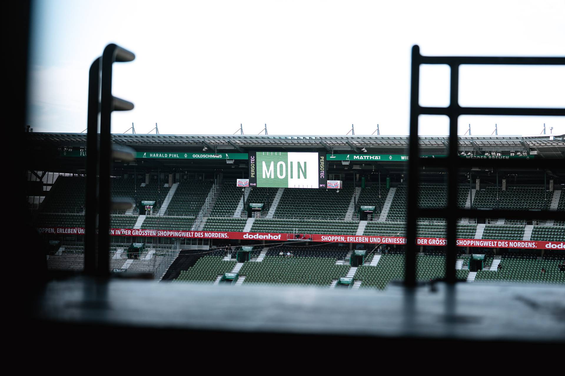Leere Stadionplätze mit einer großen digitalen Anzeige, auf der „MOIN“ zu lesen ist, gegenüber dem Aussichtspunkt, eingerahmt von dunklen Geländern.