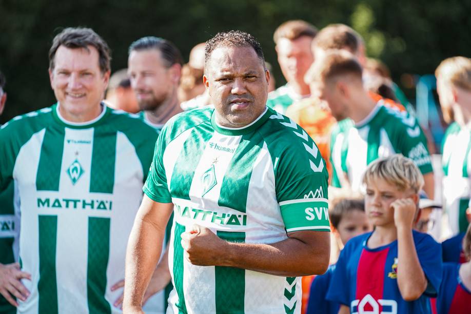 Eine Gruppe von Männern in grün-weißen Fußballtrikots steht gemeinsam im Freien, in der Nähe befinden sich Zuschauer und ein Kind in einem rot-blauen Shirt.