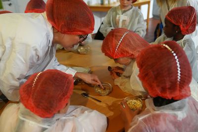 Kinder mit roten Haarnetzen und Laborkitteln untersuchen kleine Schüsseln mit Müsli an einem Tisch, angeleitet von einem Erwachsenen in ähnlicher Kleidung.