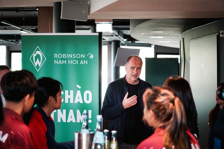 Klaus Filbry spricht zu einer Gruppe von Menschen in einem Raum mit einem Banner mit der Aufschrift „Robinson Nam Hoi An” im Hintergrund.