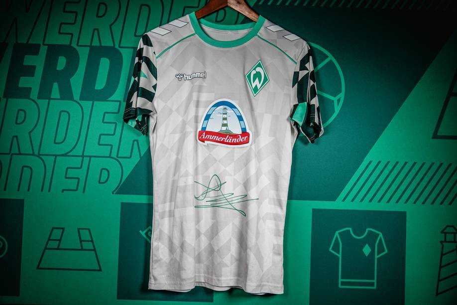 Ein grün-weißes Aufwärm-Shirt mit Unterschrift von Amira Arfaoui hängt vor einer grünen Wand mit diversen Schriftzügen und Symbolen