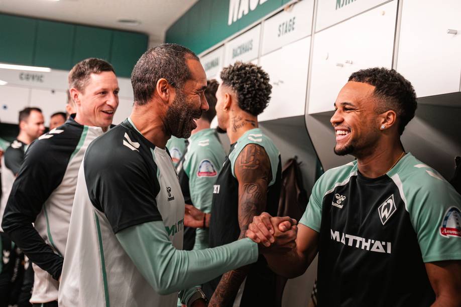 Daniel Thioune und Felix Agu klatschen in der Kabine ab. Beide lächeln sich an.