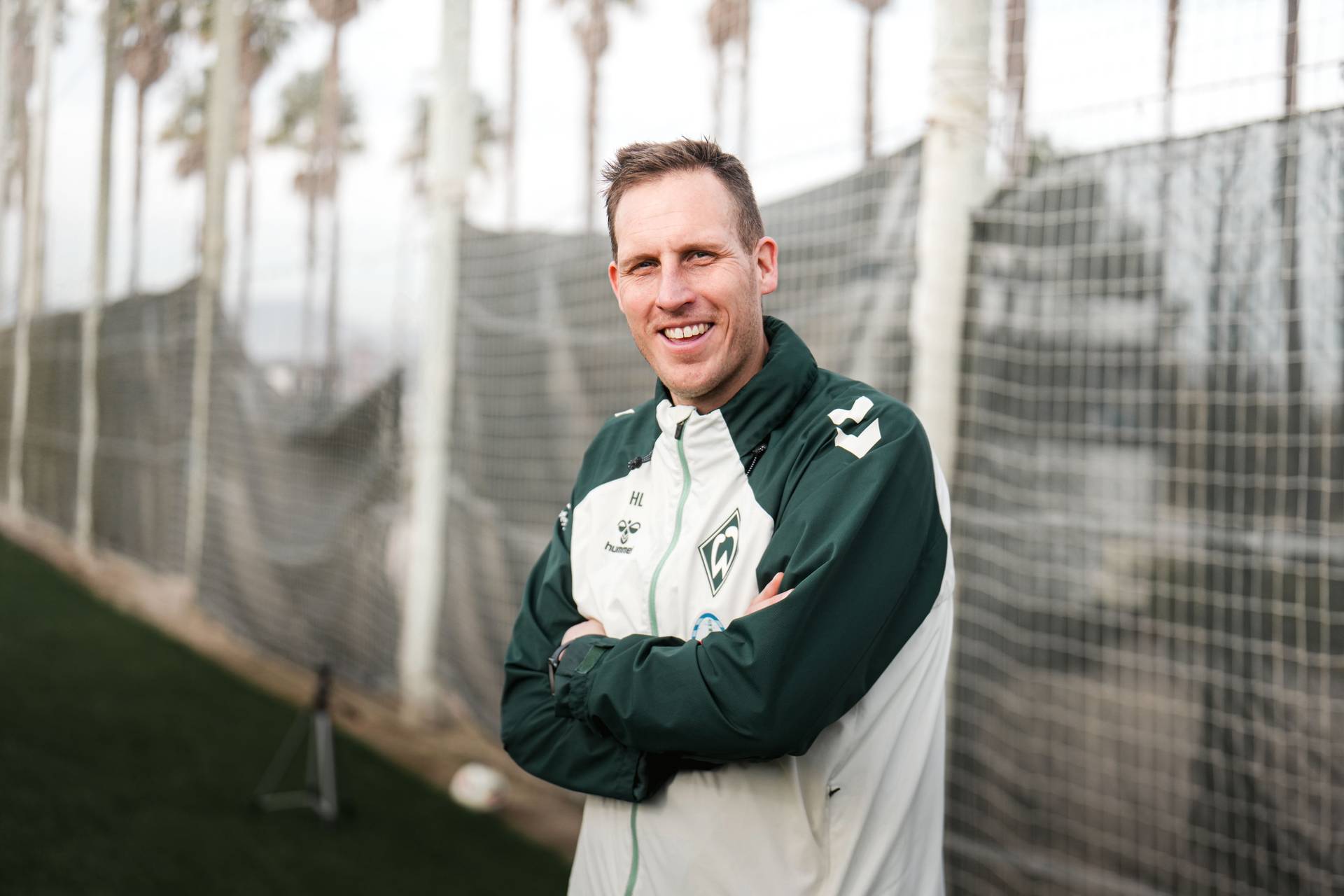 Hendrik Lemke in einer grün-weißen Sportjacke lächelt mit verschränkten Armen auf einem Fußballplatz, im Hintergrund sind ein Netz und Bäume zu sehen.