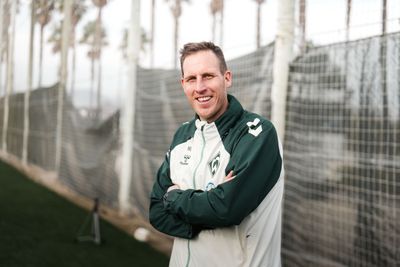Hendrik Lemke in einer grün-weißen Sportjacke lächelt mit verschränkten Armen auf einem Fußballplatz, im Hintergrund sind ein Netz und Bäume zu sehen.