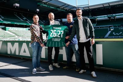 Vier Männer stehen in einem Stadion in der Nähe des Spielfelds und halten ein grünes Fußballtrikot mit der Aufschrift „WERDER 2621“ auf dem Rücken in den Händen.