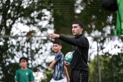 Hasan Hourou steht auf einem Bolzplatz mit Ball in der Hand und gibt mit ausgestreckter Hand Anweisungen für das Training.