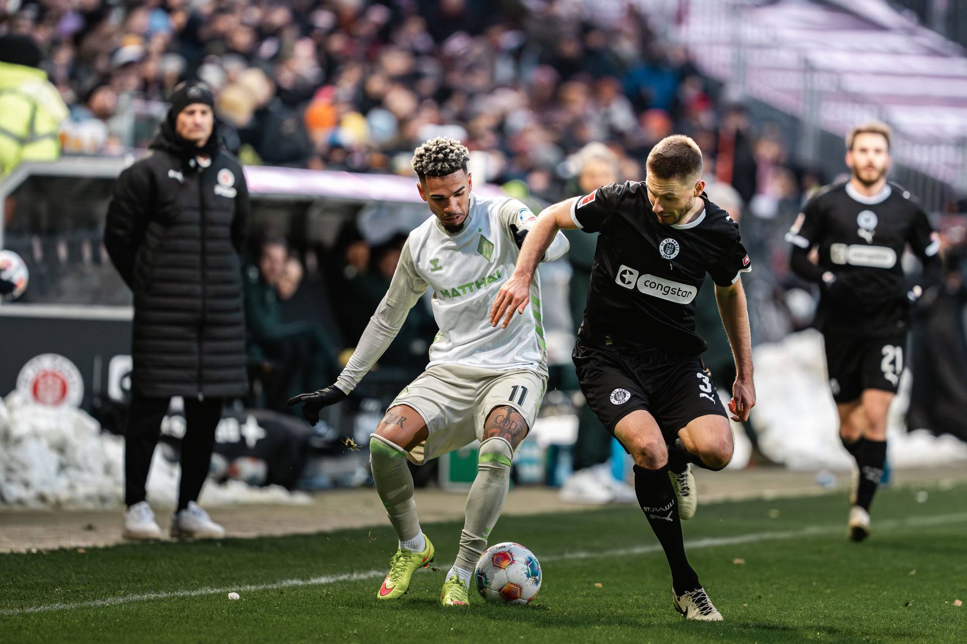 Zwei Fußballspieler kämpfen während eines Spiels auf dem Feld um den Ball, im Hintergrund sind ein Trainer und Zuschauer zu sehen.