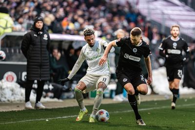 Zwei Fußballspieler kämpfen während eines Spiels auf dem Feld um den Ball, im Hintergrund sind ein Trainer und Zuschauer zu sehen.