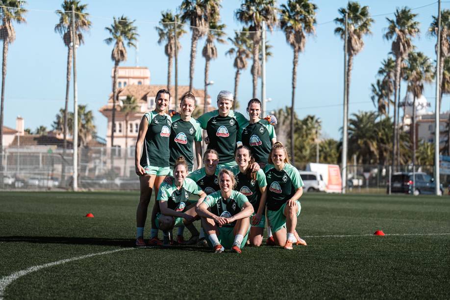 Neun Spielerinnen stehen zusammen und strahlen in die Kamera. Im Hintergrund sind Palmen und ein blauer Himmel zu sehen.