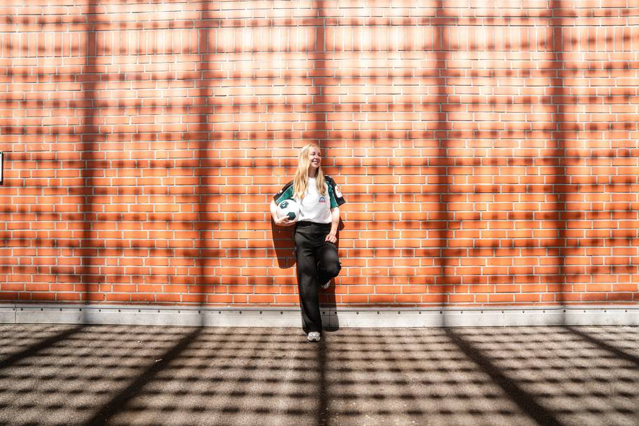 Caro Siems vor einer orangefarbenen Backsteinmauer, einen Fußball in der Hand, während karierte Schatten auf den Boden und die Mauer fallen.