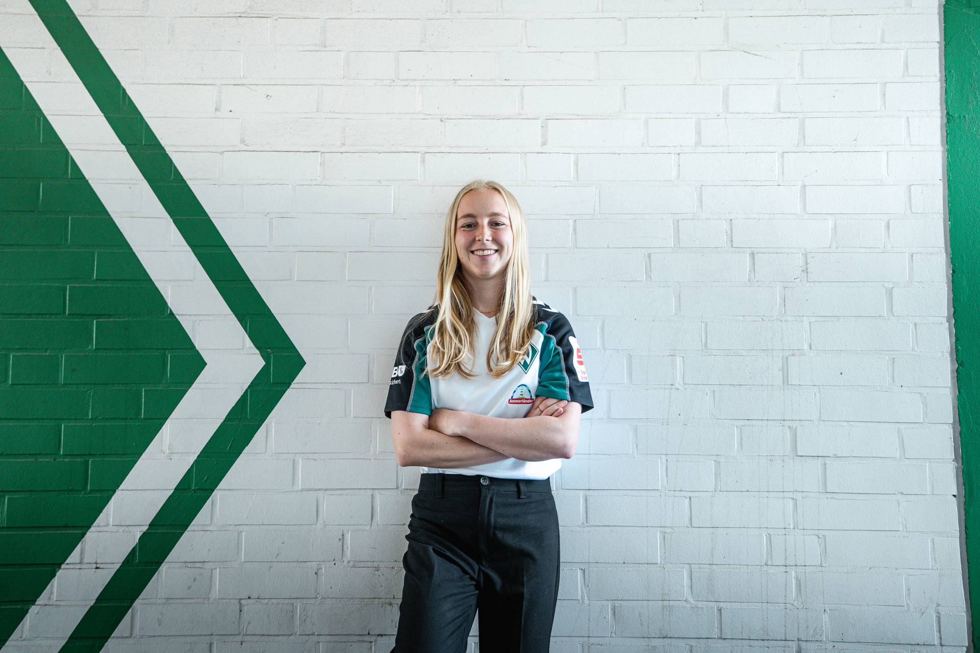 Caro Siems, die ein weiß-grünes Shirt trägt, steht lächelnd mit verschränkten Armen vor einer weißen Backsteinmauer mit einem grünen Pfeilmotiv.