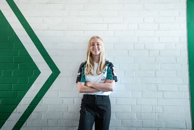 Caro Siems, die ein weiß-grünes Shirt trägt, steht lächelnd mit verschränkten Armen vor einer weißen Backsteinmauer mit einem grünen Pfeilmotiv.