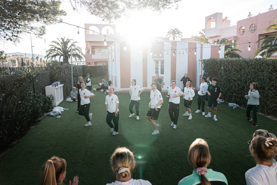 Einige Spielerinnen tanzen auf dem Rasen, im Hintergrund sind Palmen und ein Haus zu sehen.