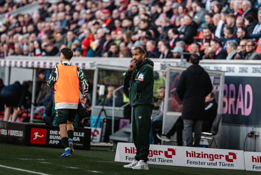 Ein Trainer steht während eines Fußballspiels nachdenklich am Spielfeldrand, während sich ein Spieler in der Nähe aufwärmt. Die Zuschauer verfolgen das Geschehen von den Tribünen aus.