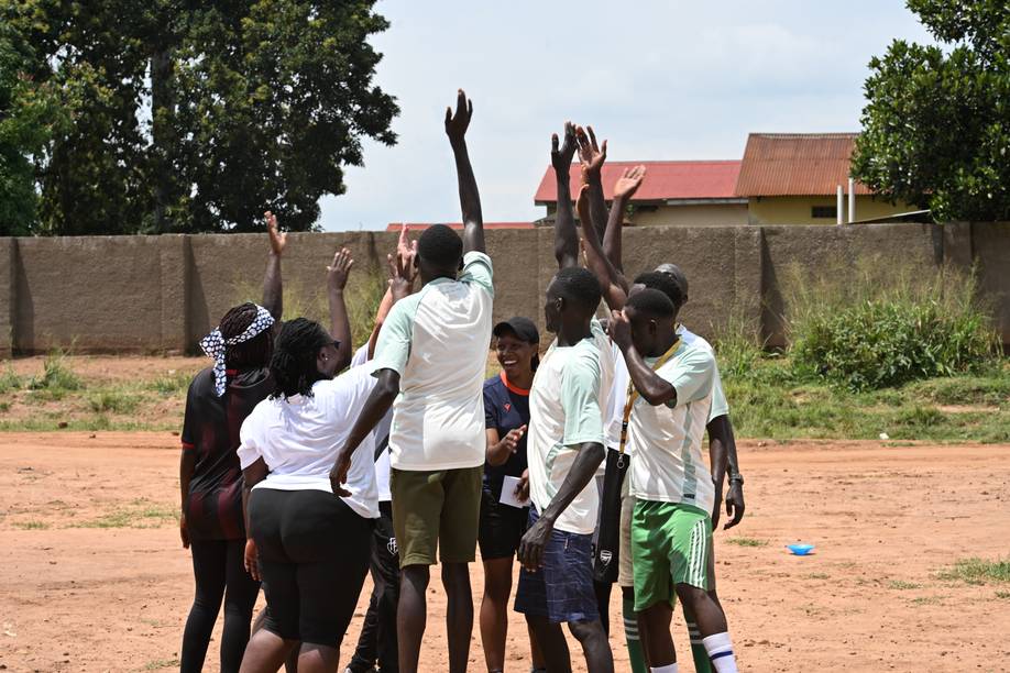 Eine Gruppe von Menschen im Freien, die gemeinsam ihre Hände in einem Kreis heben, auf einem Feld mit Bäumen und einem Gebäude im Hintergrund.