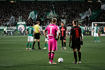 Lukas Hradecky spricht mit Robert Andrich. Beide sind von hinten zu sehen. Im Hintergrund sieht man die Fahnen in der Ostkurve.