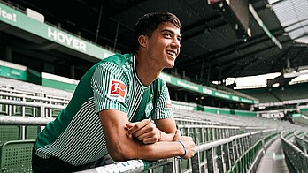 Julian Malatini lehnt über einem Wellenbrecher in der Ostkurve. Er trägt das grüne Home-Trikot.