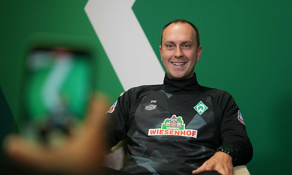 Ole Werner bei der Beantwortung der Fragen vor einem grünen Hintergrund, im Vordergrund ein Handy.