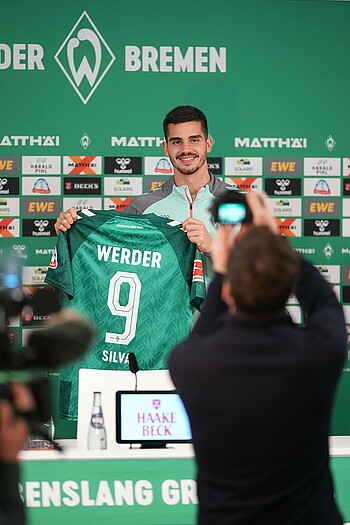 null André Silva hält ein Werder-Trikot in der Mixed Zone auf dem Podium in die Kamera