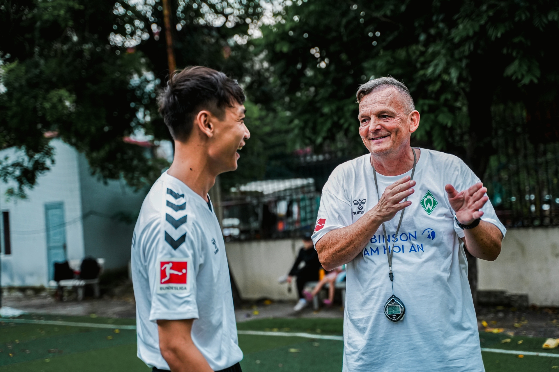 Zwei Männer in Werdertrikots stehen auf einem Fußballplatz und lachen