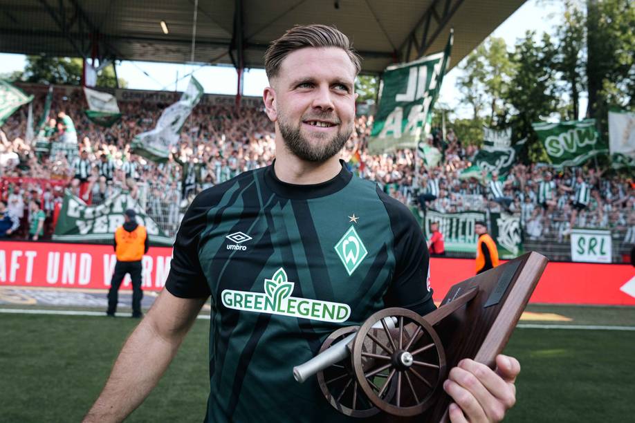 Ein Fußballspieler in einem grünen Trikot, der einen Pokal mit Kanonenmotiv in den Händen hält und auf einem Spielfeld steht, im Hintergrund jubelnde Fans und Fahnen.